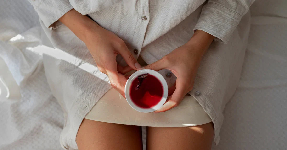 Una persona sentada sobre una cama con sábanas blancas sostiene una taza de té rojo con ambas manos, vestida con una camisa larga de color claro. La imagen está tomada desde arriba, capturando un momento de calma y calidez.